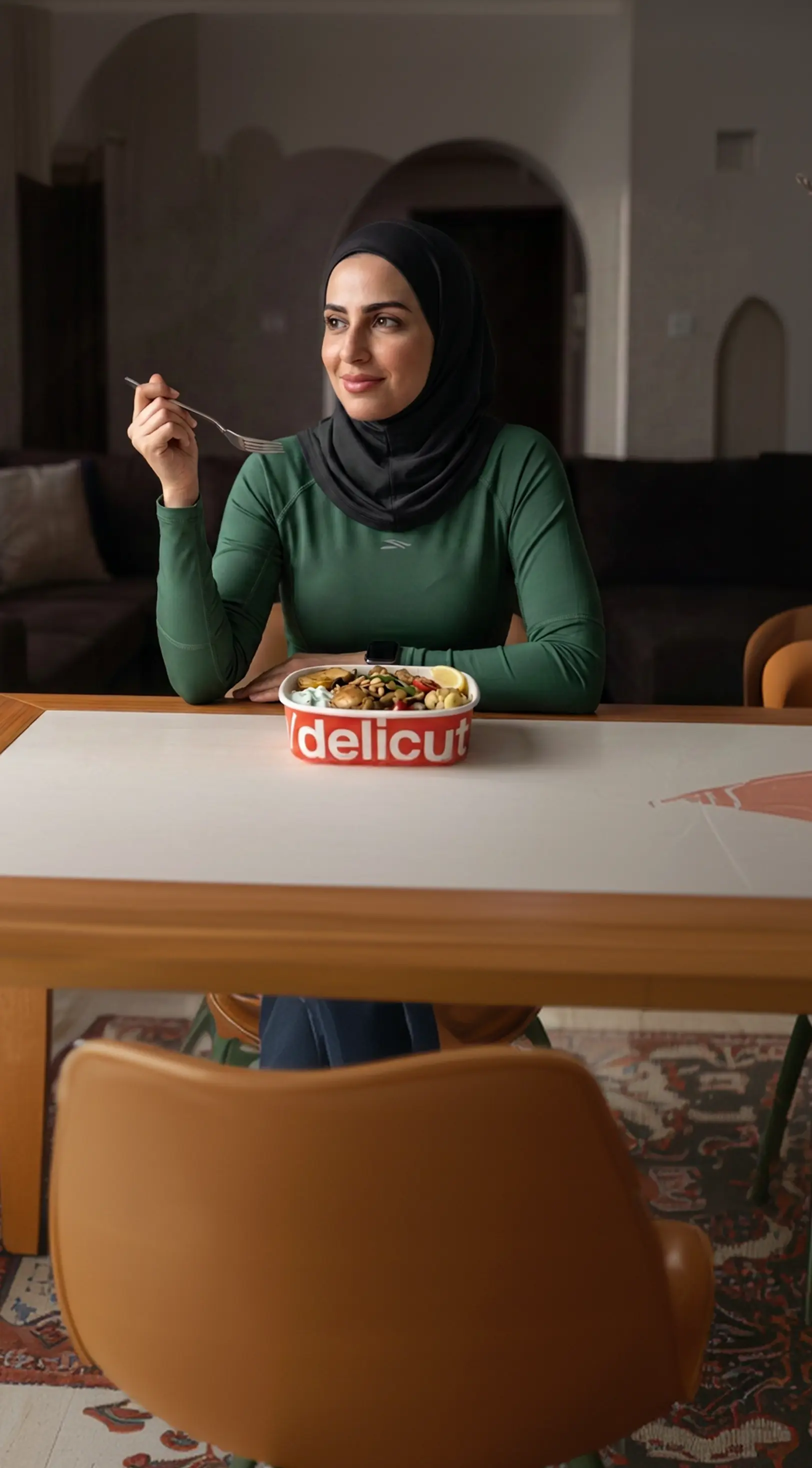 Woman in a green abaya enjoying a Delicut Arabic dish from a takeaway bowl at home